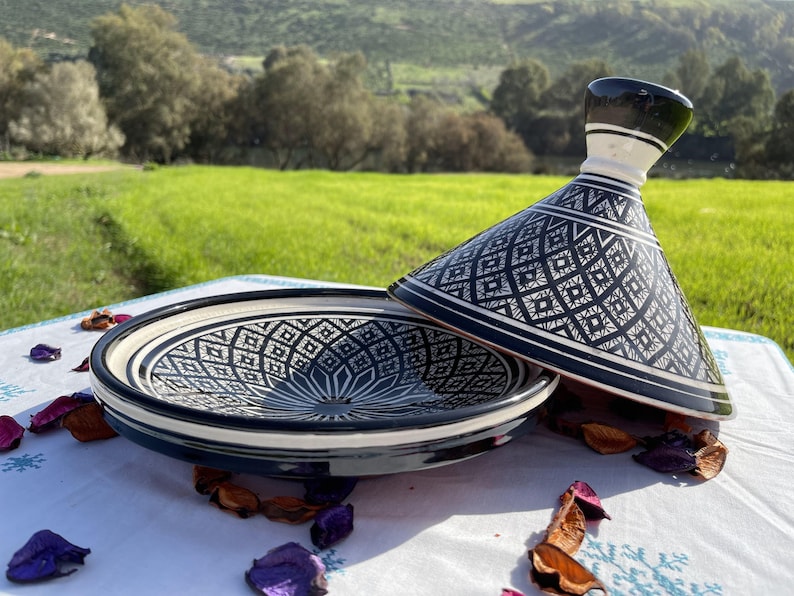 Może przedstawiać: Czarno-biała ceramiczna tagine z geometrycznym wzorem. Tagine wykonana jest z glazurowanej ceramiki i ma stożkowy pokrywę. Tagine stoi na białym obrusie z płatkami kwiat&oacute;w.