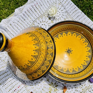 May include: A yellow ceramic tagine with a black geometric design. The tagine has a lid and a base. The lid is round and has a small spout. The base is round and has a wide opening.
