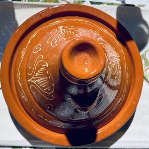 May include: A terracotta tagine with a lid featuring a decorative design of white swirls and lines. The lid has a small spout for steam release.