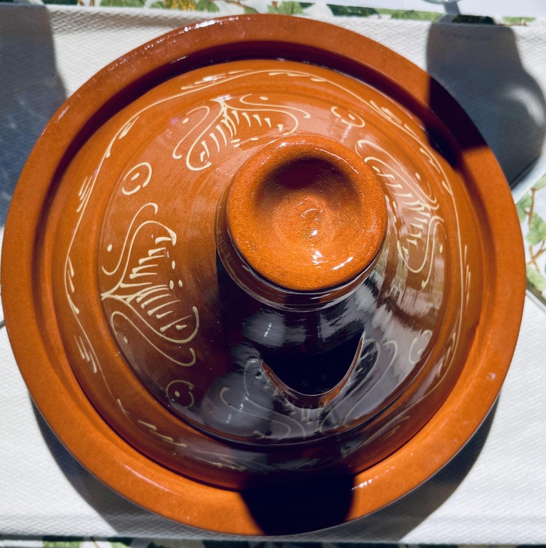 May include: A terracotta tagine with a lid featuring a decorative design of white swirls and lines. The lid has a small spout for steam release.
