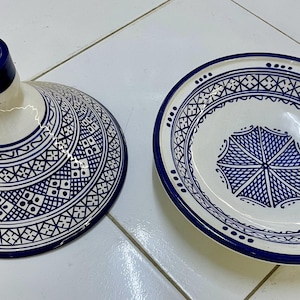 May include: A white ceramic tagine with a blue and white geometric pattern. The tagine has a round base and a conical lid with a small opening at the top.