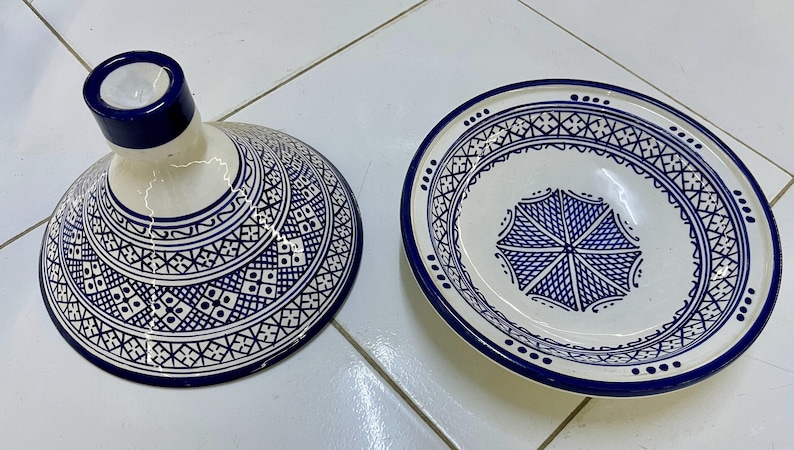May include: A white ceramic tagine with a blue and white geometric pattern. The tagine has a round base and a conical lid with a small opening at the top.