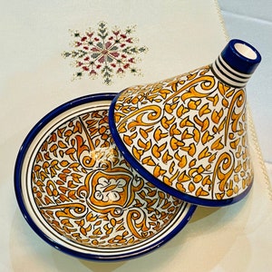 May include: A white ceramic tagine with a blue rim and intricate yellow and orange floral patterns. The tagine is open, revealing the matching bowl underneath.