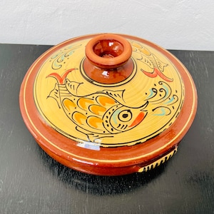 May include: A terracotta ceramic serving dish with a lid, featuring a hand-painted design of two stylized fish in yellow, orange, blue, and red on a cream background.