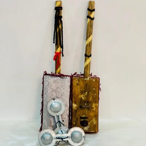 May include: Two wooden musical instruments with strings, called a kora, with a red and white patterned fabric covering the sound box. The instruments are standing upright on a white surface. There are three silver jingle bells in the foreground.
