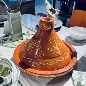 May include: A brown ceramic tagine with a white design. The tagine is on a white table with a white tablecloth and green napkins.