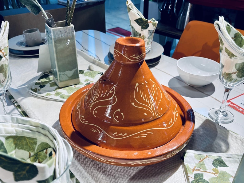 May include: A brown ceramic tagine with a white design. The tagine is on a white table with a white tablecloth and green napkins.