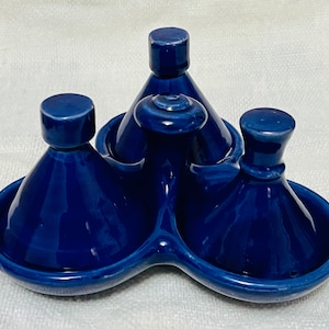 Puede incluir: Un juego de condimentos de cerámica azul cobalto. El juego incluye tres recipientes individuales con cuerpos cónicos y tapas redondeadas, colocados en una bandeja conectada. La bandeja tiene una forma curva única. El juego está diseñado para servir condimentos.