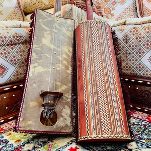 May include: Two traditional Moroccan musical instruments, a  tanbura and a gimbri, with intricate wood carvings and decorative details. The tanbura has a light brown, leather-covered body with a wooden neck and strings. The gimbri has a dark brown, wooden body with a decorative inlay of wood and bone. Both instruments are resting on a colorful rug.