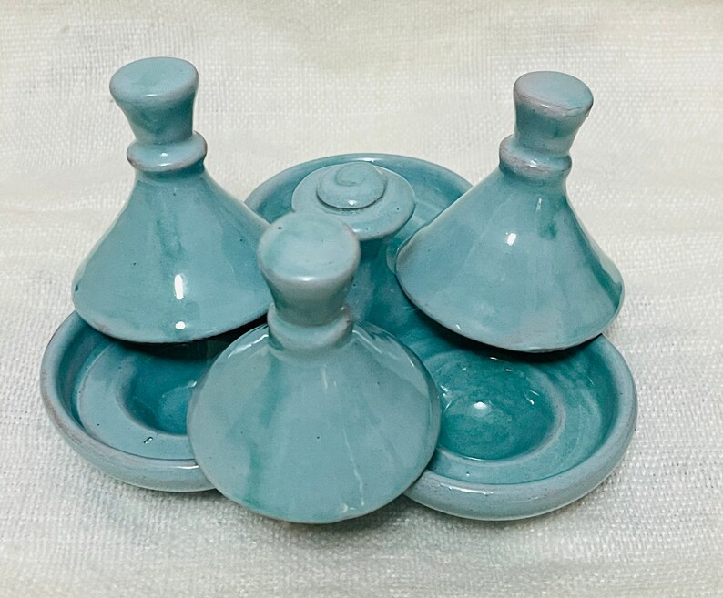 May include: A set of four teal ceramic condiment dishes with lids. The dishes are arranged in a circle with the lids placed on top of the dishes.