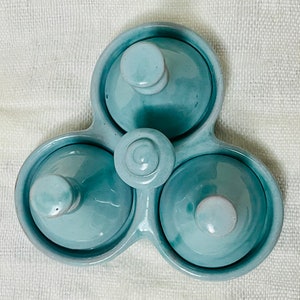 May include: A light blue ceramic three-section dish with three individual lids. Each lid has a small knob on top. The dish is designed to hold small items, such as spices or condiments.