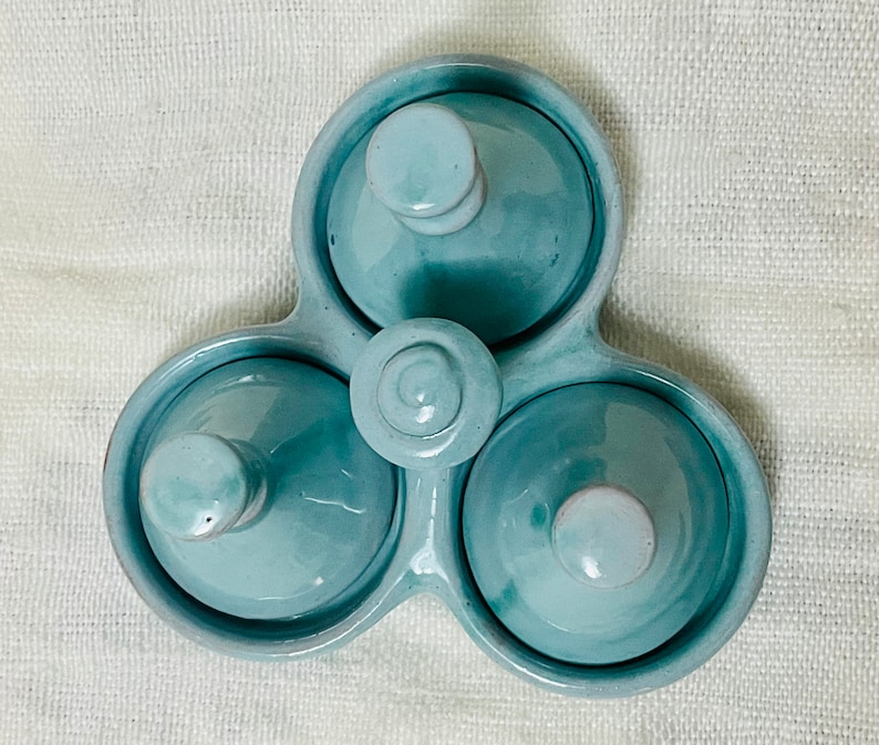 May include: A light blue ceramic three-section dish with three individual lids. Each lid has a small knob on top. The dish is designed to hold small items, such as spices or condiments.