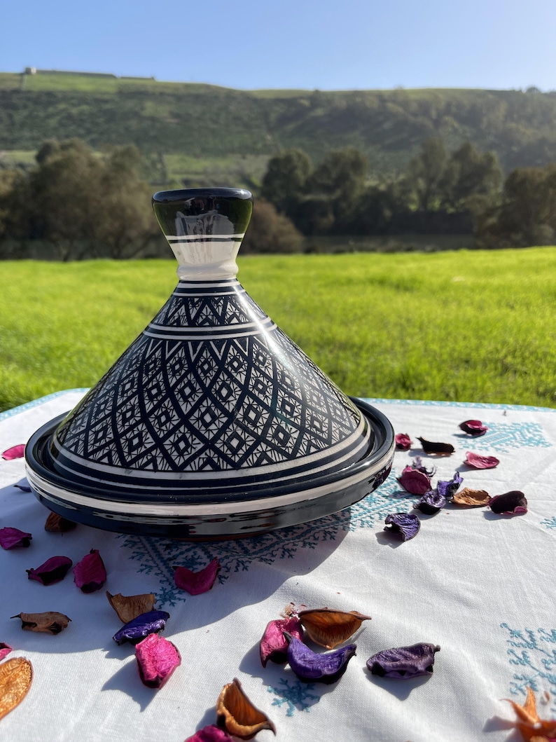 Może przedstawiać: Czarno-biała ceramiczna pokrywa do tagine z misternymi geometrycznymi wzorami. Pokrywa leży na białym obrusie z rozrzuconymi płatkami kwiat&oacute;w.