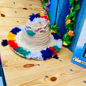 May include: A straw hat with a wide brim and colorful tassels around the edge. The hat is made of woven straw and has a white and pink pattern on the crown.