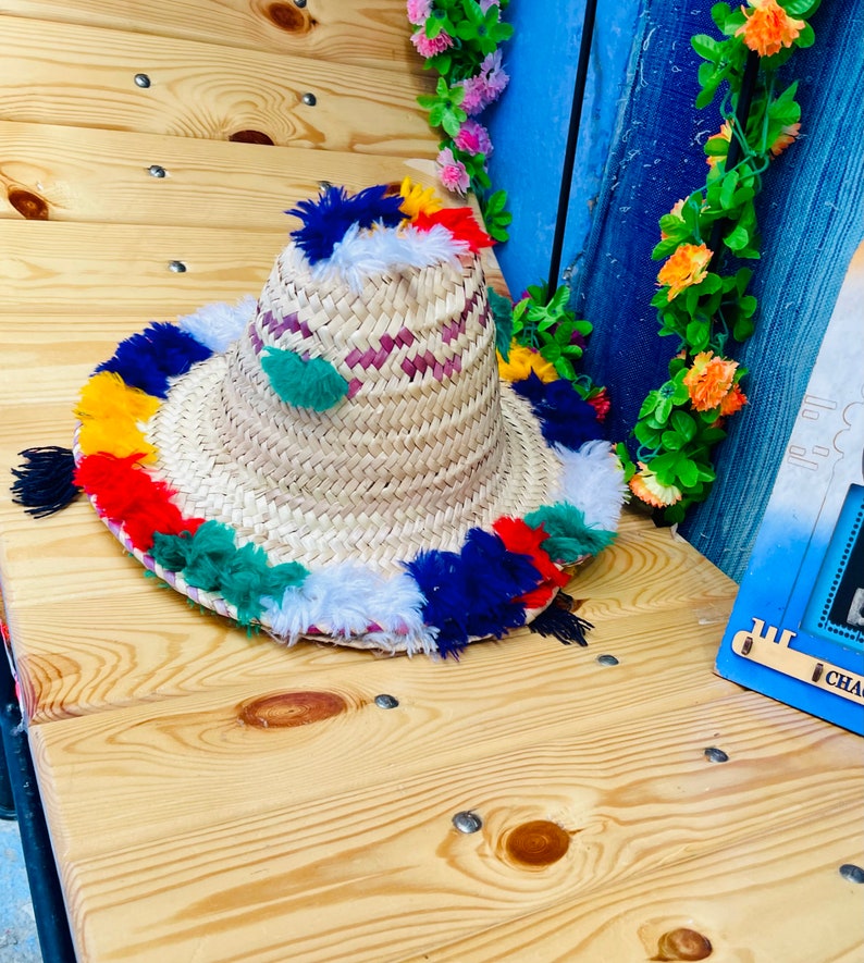 May include: A straw hat with a wide brim and colorful tassels around the edge. The hat is made of woven straw and has a white and pink pattern on the crown.