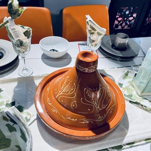 May include: A terracotta tagine with a white floral design, set on a white tablecloth with green and white napkins. The tagine is surrounded by plates, glasses, and vases.