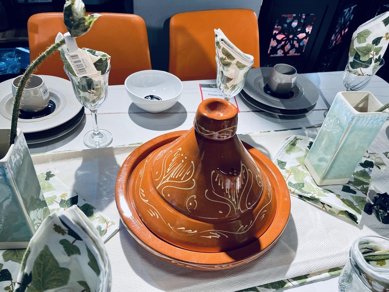 May include: A terracotta tagine with a white floral design, set on a white tablecloth with green and white napkins. The tagine is surrounded by plates, glasses, and vases.