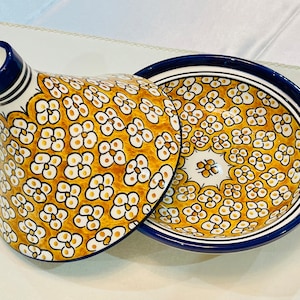 May include: A ceramic tagine with a conical lid and a matching base. The tagine is decorated with a repeating floral pattern in yellow, white, and brown against a yellow background. The rim and spout are blue.