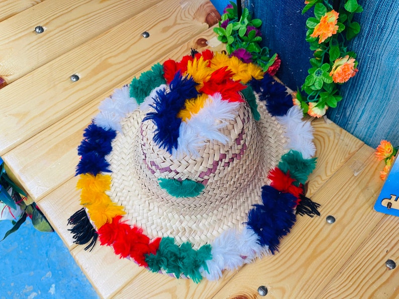 May include: A straw hat with colorful pom-poms around the brim. The pom-poms are red, yellow, green, blue, and white.