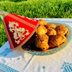 May include: A red and gold embroidered hat sits on top of a golden tray filled with freshly baked croissants. The tray is on a white tablecloth with a blue embroidered pattern.