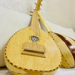 Könnte beinhalten: Zwei Ouds aus Holz, traditionelle Saiteninstrumente aus dem Nahen Osten, werden gezeigt. Die vordere Oud hat einen hellen Holzkörper mit einem dekorativen Schallloch und einem Steg. Die zweite Oud hat ein gestreiftes Design in Braun- und Beigetönen.