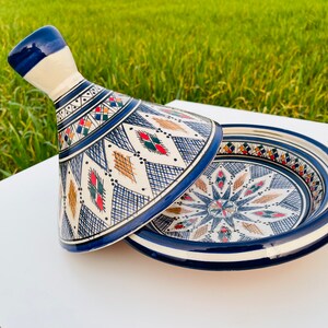 Puede incluir: Una tagine de cerámica blanca con un patrón geométrico azul y blanco. La tapa está decorada con un patrón geométrico azul, blanco, rojo y verde. La tagine está sobre una superficie blanca.