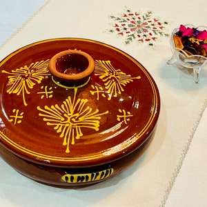 May include: A brown ceramic casserole dish with a lid. The dish has a yellow floral design and a small knob on the lid.