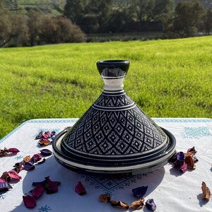 Może przedstawiać: Czarno-biała ceramiczna tagine z geometrycznym wzorem. Tagine stoi na białym obrusie z r&oacute;żowymi i fioletowymi płatkami kwiat&oacute;w.
