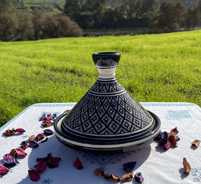 Może przedstawiać: Czarno-biała ceramiczna tagine z geometrycznym wzorem. Tagine stoi na białym obrusie z r&oacute;żowymi i fioletowymi płatkami kwiat&oacute;w.