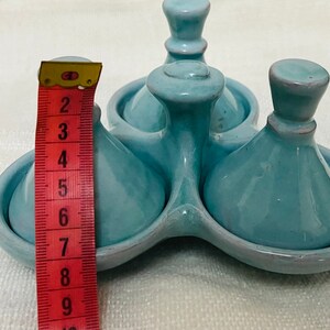 May include: A set of three blue ceramic cruets with a matching blue ceramic base. The cruets have a rounded, bulbous shape with a small spout at the top. The base has three indentations to hold the cruets.