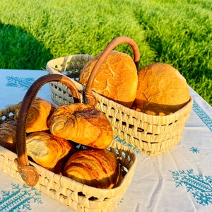 May include: Two woven baskets filled with baked goods sit on a white tablecloth with a blue pattern. One basket holds croissants, the other holds round loaves of bread. Both baskets have brown leather handles. The background is a green field.