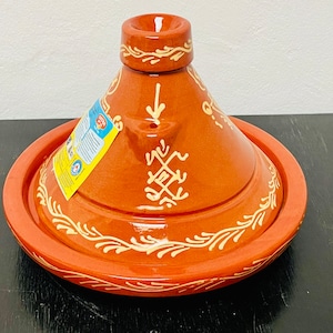 May include: A terracotta tagine with a white glaze and intricate designs. The tagine has a round base and a conical lid with a small opening at the top. The tagine is a traditional Moroccan cooking pot.