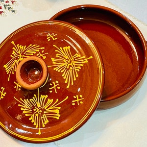 May include: A brown ceramic casserole dish with a lid. The lid features yellow floral designs and a small handle. The dish is open, revealing its interior. The design is rustic and handmade.