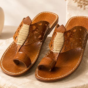 May include: A pair of tan leather sandals featuring a toe loop design, intricate laser-cut floral patterns on the wide strap, and a central beige suede accent. The footwear includes visible white stitching along the edges of the soles.