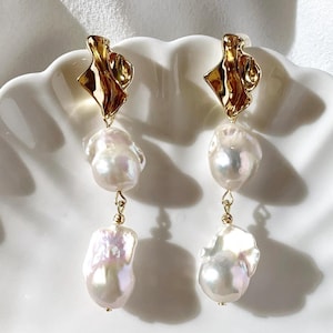 Puede incluir: Un par de elegantes pendientes con grandes perlas blancas irregulares suspendidas de la parte superior dorada de forma abstracta. Las perlas tienen un brillo lustroso, y los pendientes se muestran en un plato blanco festoneado.