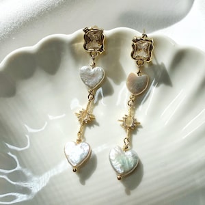 May include: A pair of gold-tone drop earrings featuring a rectangular clear crystal stud, two white heart-shaped iridescent beads, and a central star-shaped charm with small clear stones. The earrings are constructed with delicate gold-tone metal chains.