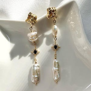 May include: A pair of gold-toned drop earrings featuring a textured gold stud, a white baroque pearl, and a central gold star-shaped charm with a small black stone. The earrings finish with a long, irregular white pearl dangling at the bottom.