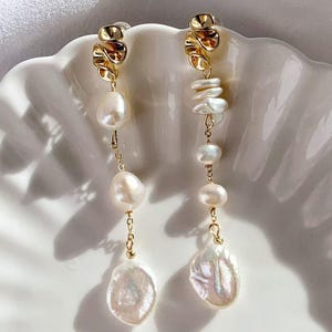 Puede incluir: Un par de pendientes de oro con gotas de perlas blancas. Los pendientes presentan un diseño de espiral dorado en la parte superior y una perla grande de forma irregular en la parte inferior.