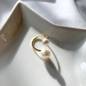 K&ouml;nnte beinhalten: Ein goldfarbener Ring mit zwei wei&szlig;en Perlen. Der Ring hat ein schlichtes, offenes Design, mit einer Perle oben und einer unten. Der Ring wird auf einer wei&szlig;en Oberfl&auml;che pr&auml;sentiert.