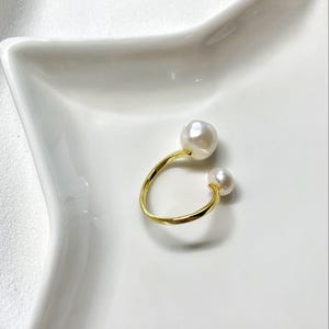 K&ouml;nnte beinhalten: Ein goldfarbener Ring mit zwei wei&szlig;en Perlen. Der Ring hat ein offenes Design, mit einer gro&szlig;en Perle oben und einer kleineren Perle unten. Der Ring ist vor einem wei&szlig;en Hintergrund platziert.