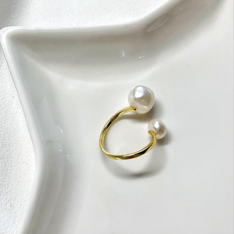 K&ouml;nnte beinhalten: Ein goldfarbener Ring mit zwei wei&szlig;en Perlen. Der Ring hat ein offenes Design, mit einer gro&szlig;en Perle oben und einer kleineren Perle unten. Der Ring ist vor einem wei&szlig;en Hintergrund platziert.