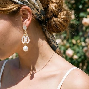 May include: A gold-tone drop earring featuring a white teardrop-shaped stud, a central oval loop composed of small white seed pearls, and a single large white pearl dangling at the bottom, suspended from a gold-tone metal chain link.