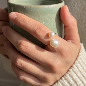 K&ouml;nnte beinhalten: Ein goldener Ring mit zwei wei&szlig;en, gl&auml;nzenden Perlen. Der Ring befindet sich an einem Finger, mit einer hellgr&uuml;nen Tasse im Hintergrund. Die Person tr&auml;gt einen cremefarbenen Pullover.