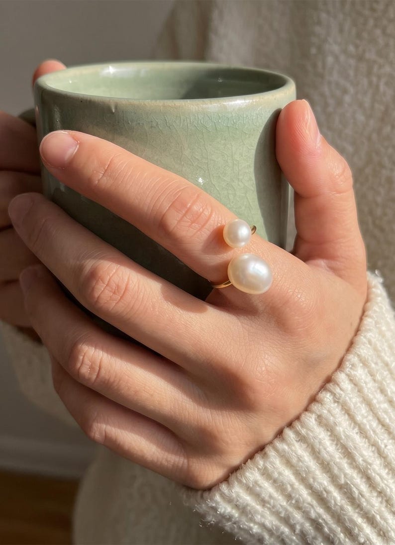 K&ouml;nnte beinhalten: Ein goldener Ring mit zwei wei&szlig;en, gl&auml;nzenden Perlen. Der Ring befindet sich an einem Finger, mit einer hellgr&uuml;nen Tasse im Hintergrund. Die Person tr&auml;gt einen cremefarbenen Pullover.