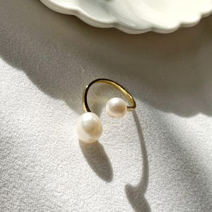K&ouml;nnte beinhalten: Ein goldfarbener Ring mit zwei wei&szlig;en Perlen. Eine Perle ist gr&ouml;&szlig;er und unregelm&auml;&szlig;ig geformt, die andere kleiner und rund. Der Ring hat ein minimalistisches Design mit einem gebogenen Band, das die Perlen h&auml;lt.