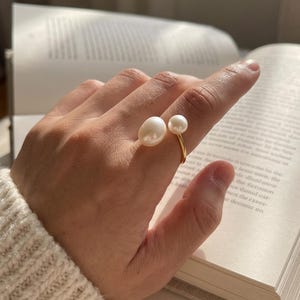 K&ouml;nnte beinhalten: Ein goldener Ring mit zwei wei&szlig;en Perlen. Eine Perle ist oval und gr&ouml;&szlig;er, die andere rund und kleiner. Der Ring wird an einem Finger getragen, im Hintergrund ein offenes Buch.