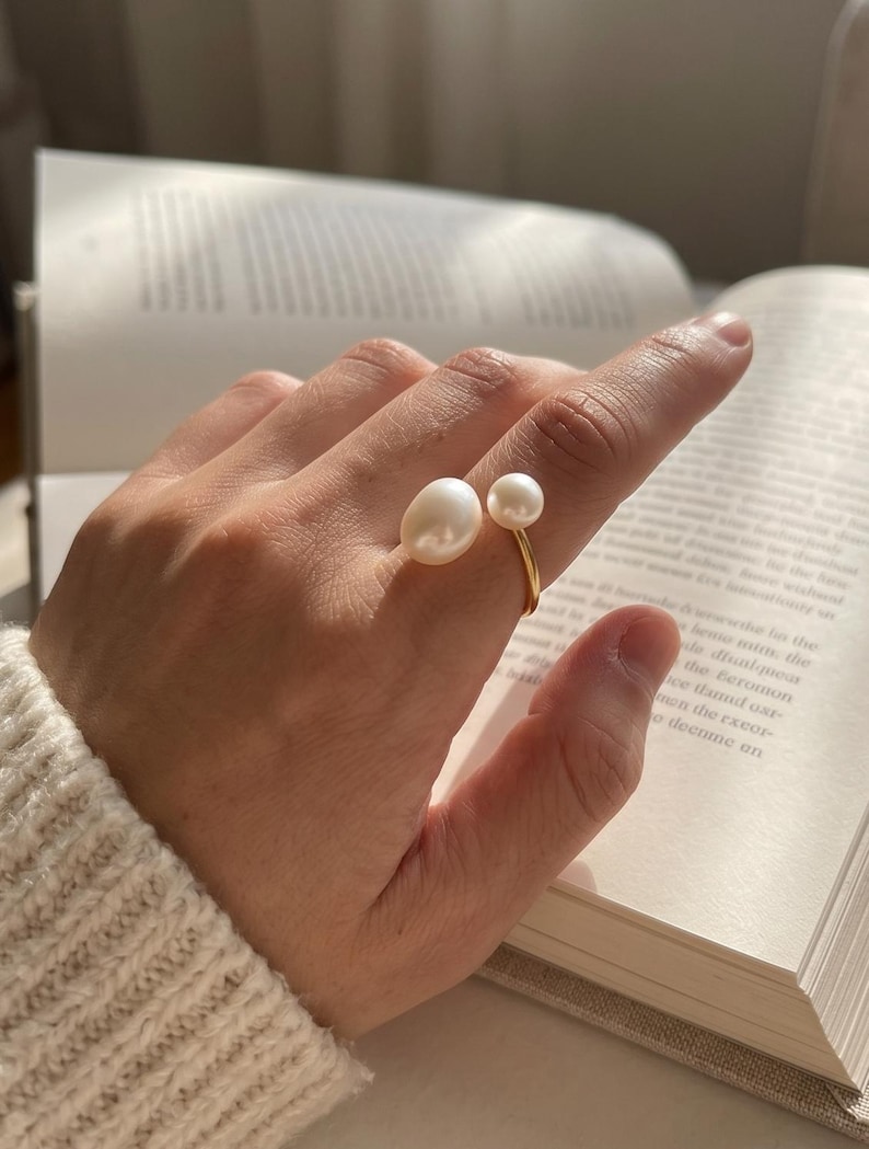 K&ouml;nnte beinhalten: Ein goldener Ring mit zwei wei&szlig;en Perlen. Eine Perle ist oval und gr&ouml;&szlig;er, die andere rund und kleiner. Der Ring wird an einem Finger getragen, im Hintergrund ein offenes Buch.