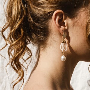 May include: A gold-toned drop earring featuring a small circular stud, a central section composed of three delicate loops of small white seed pearls, and a single larger, irregular white baroque pearl suspended at the very bottom of the piece.