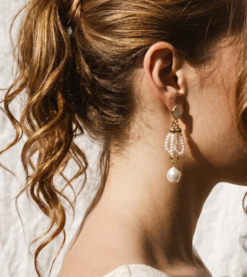 May include: A gold-toned drop earring featuring a small circular stud, a central section composed of three delicate loops of small white seed pearls, and a single larger, irregular white baroque pearl suspended at the very bottom of the piece.