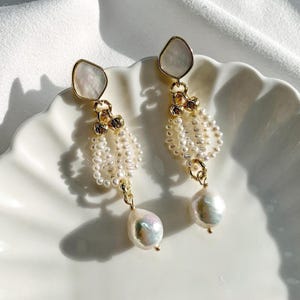 May include: A pair of gold-toned drop earrings featuring a top irregular-shaped white shell component, a central section composed of multiple strands of small white seed pearls, and a single large, lustrous white baroque pearl at the base.